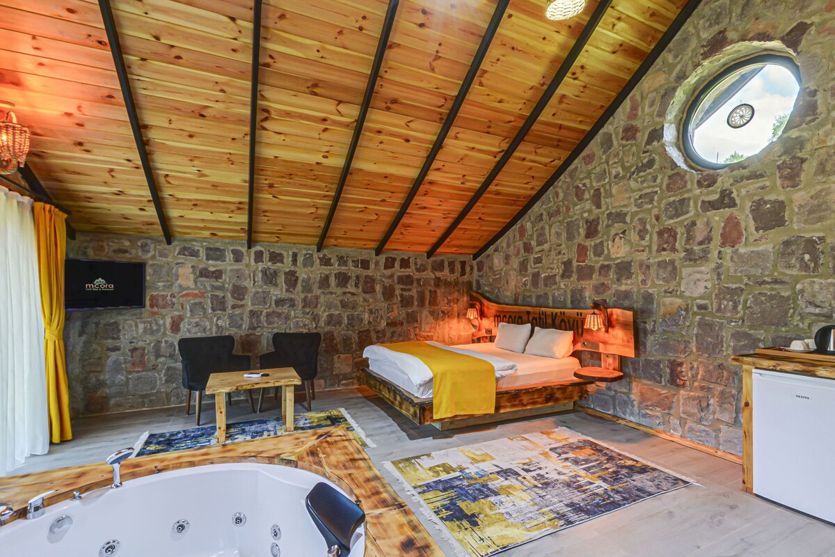 A room with a stone wall, wooden ceiling, a bed, a small table, a chair, a bathtub, and a television.