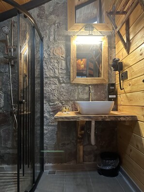 A bathroom with a stone wall, a wooden vanity, a white sink, a glass-enclosed shower, and a wooden ceiling.