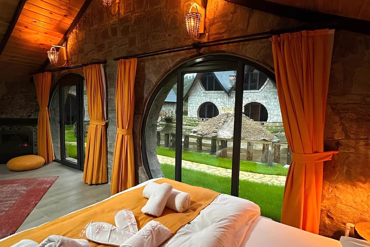 A bedroom with a large bed, a fireplace, and a view of a stone building through large windows.