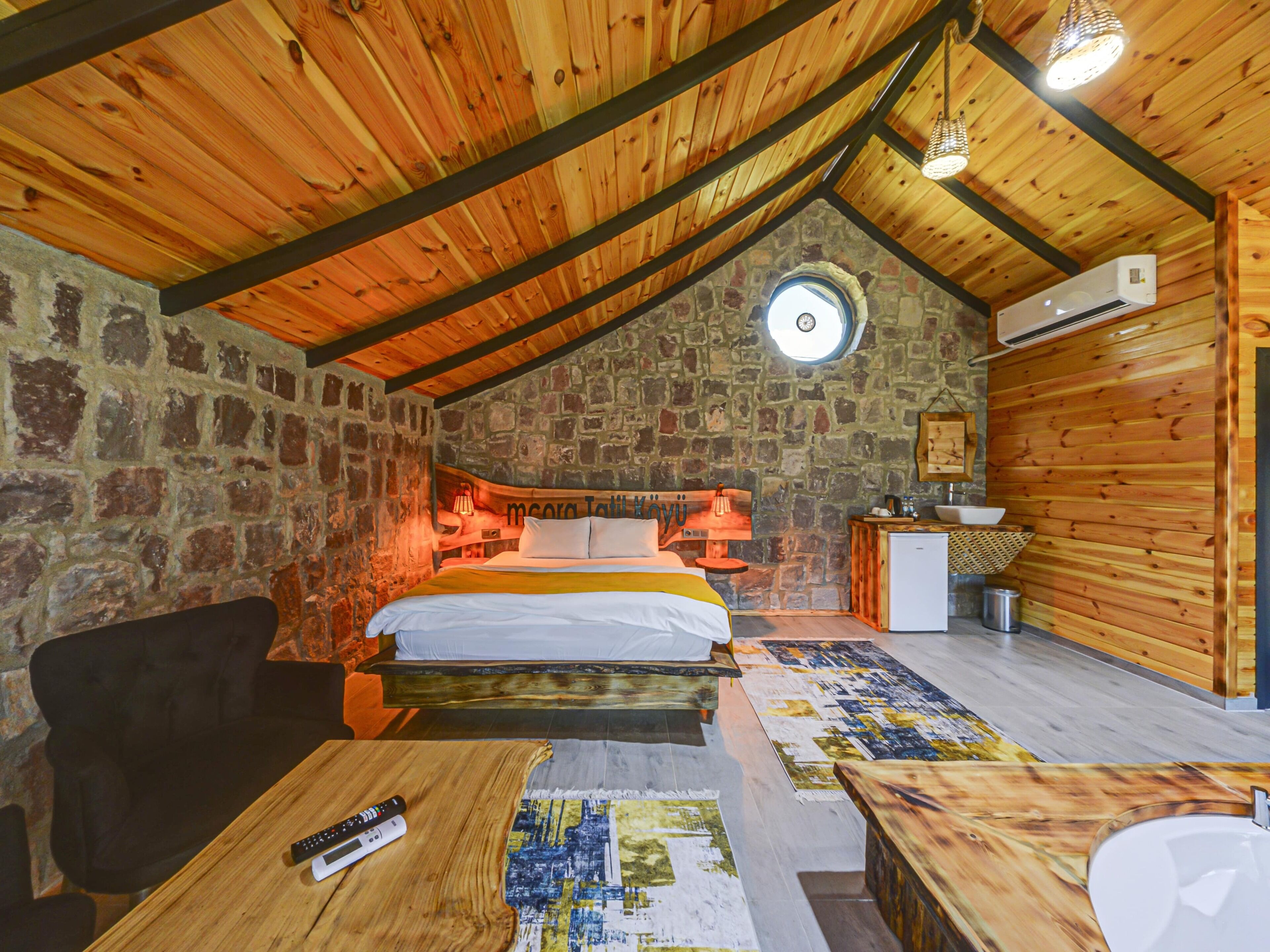 Ein gemütliches Schlafzimmer unter dem Dach mit einer Steinwand, Holzbalkendecke, einem Bett, einem Schreibtisch und einer kleinen Küchenzeile.