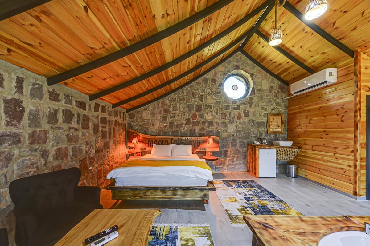 A cozy attic bedroom with a stone wall, wooden ceiling beams, a bed, a desk, and a small kitchenette.