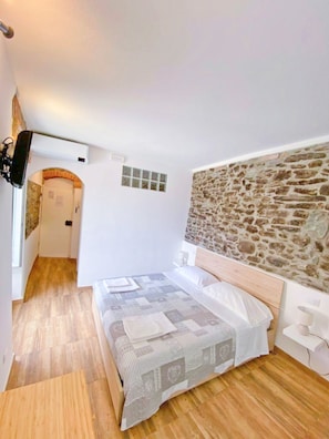 A bedroom with a stone wall headboard, a wooden floor, a bed with a patterned bedspread, and a TV mounted on the wall. - Double room "Malvasia" - Casedirio (Riomaggiore)