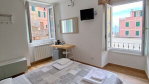 Een slaapkamer met een bed, een bureau, een stoel, een televisie en grote ramen met uitzicht op gebouwen.