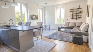 Une cuisine moderne dotée d’un îlot central, un coin repas avec une table et des chaises en verre, un espace salon confortable avec un canapé et un pouf, et de grandes fenêtres qui laissent entrer la lumière naturelle.