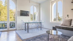 Un salon moderne avec une table à manger en verre, des chaises transparentes, un canapé gris et de grandes fenêtres donnant sur la verdure.