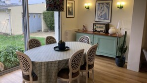 A dining area with a round table, six chairs, a patterned tablecloth, a hanging lamp, a kitchen area with a vintage radio, and a cactus plant. - Townhome (Lanester)