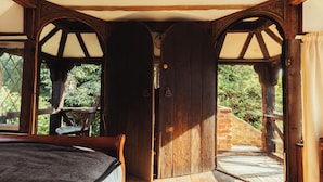 A rustic wooden room with a bed, a bench, and a view of greenery outside.