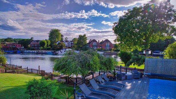 A pool area with lounge chairs, a wooden deck, and a view of a river with boats and houses in the background.