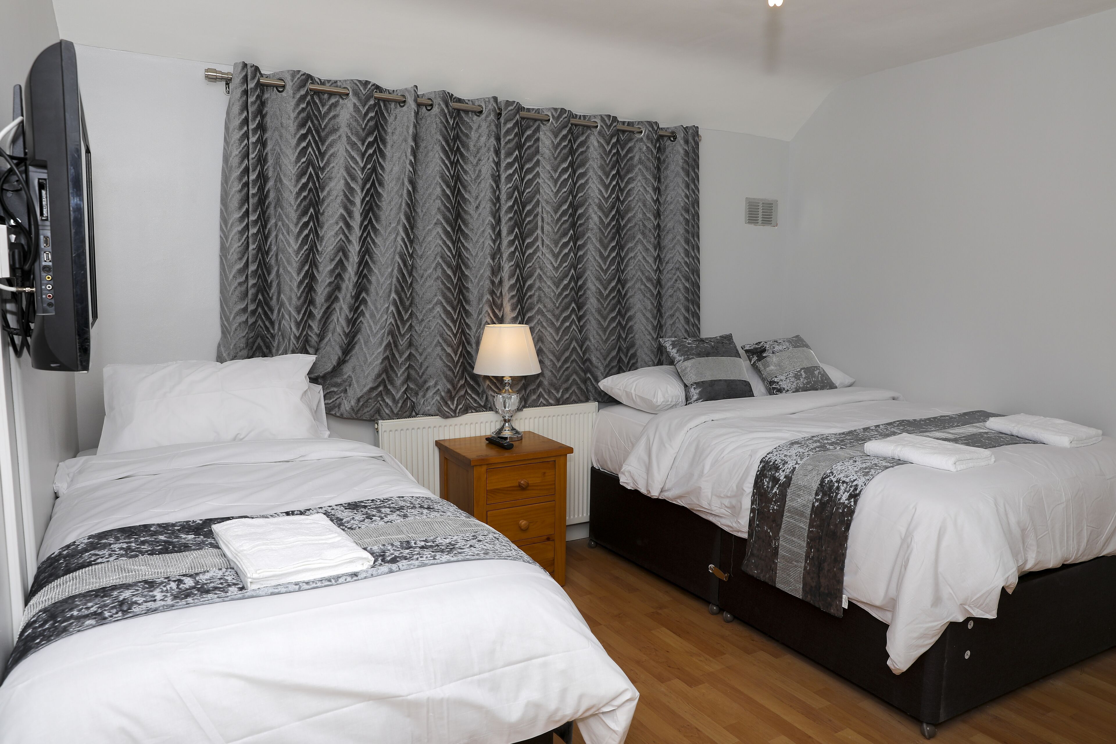 A bedroom with two beds, a nightstand, a lamp, and a TV mounted on the wall.