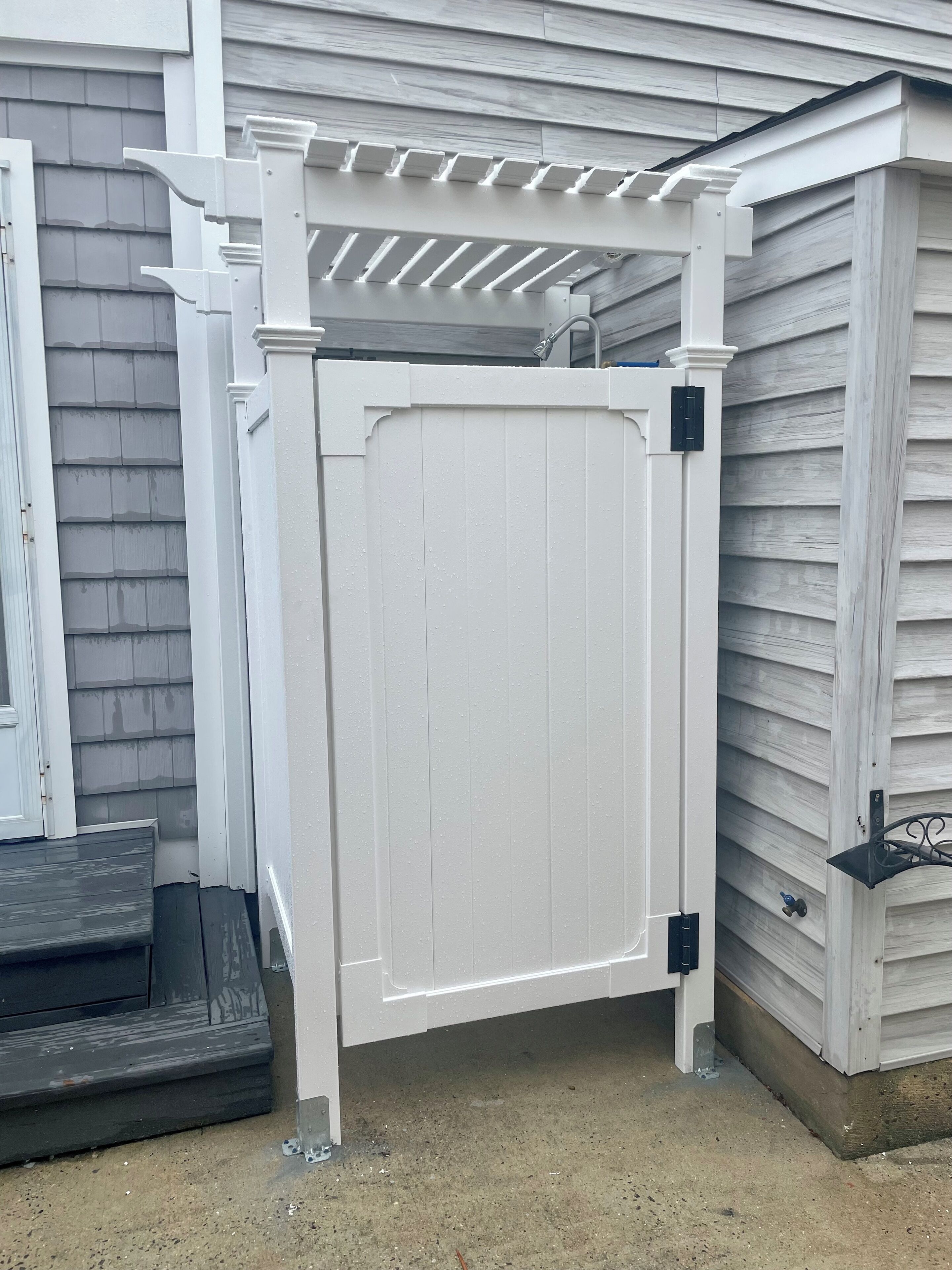A white wooden gate with a metal latch, attached to a white wooden pergola with a roof, set against a house with gray siding.