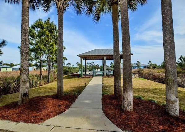 A gazebo with a concrete pathway leading to it, surrounded by palm trees and a grassy area.
