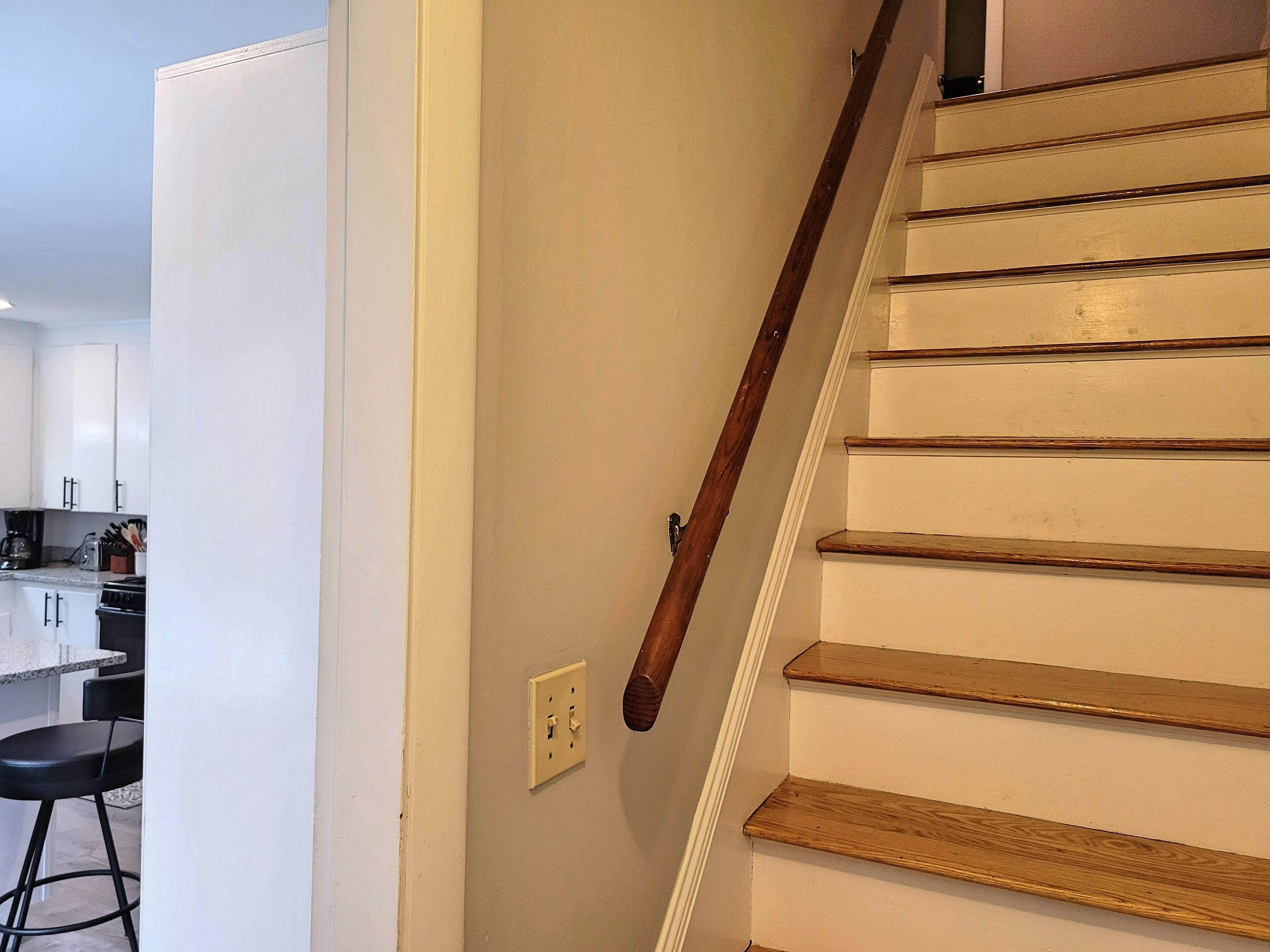 Una escalera de madera con barandilla que lleva a una zona de cocina con armarios blancos y un taburete de bar.