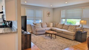 A modern living room with a sectional sofa, a coffee table, and a TV. There are windows with blinds, a kitchen area with a stove and kettle, and a rug on the floor.