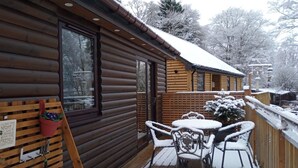 A wooden cabin with a deck, a table, and chairs covered in snow.