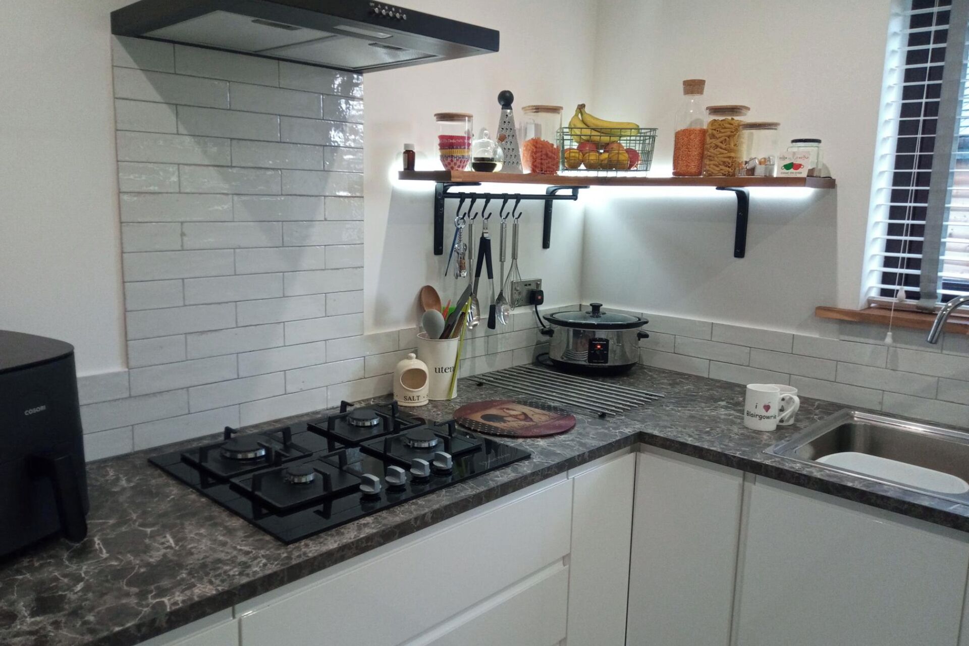 Une cuisine équipée d’une cuisinière à gaz, d’un autocuiseur et d’un évier. On y trouve des étagères remplies de bocaux et une fenêtre avec des stores.