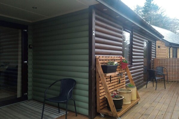 A log cabin with a green exterior, a wooden porch with a planter, and a chair. - 2 Bed Cottage Sleeps 4 -private Garden -pets Okay (Blairgowrie)