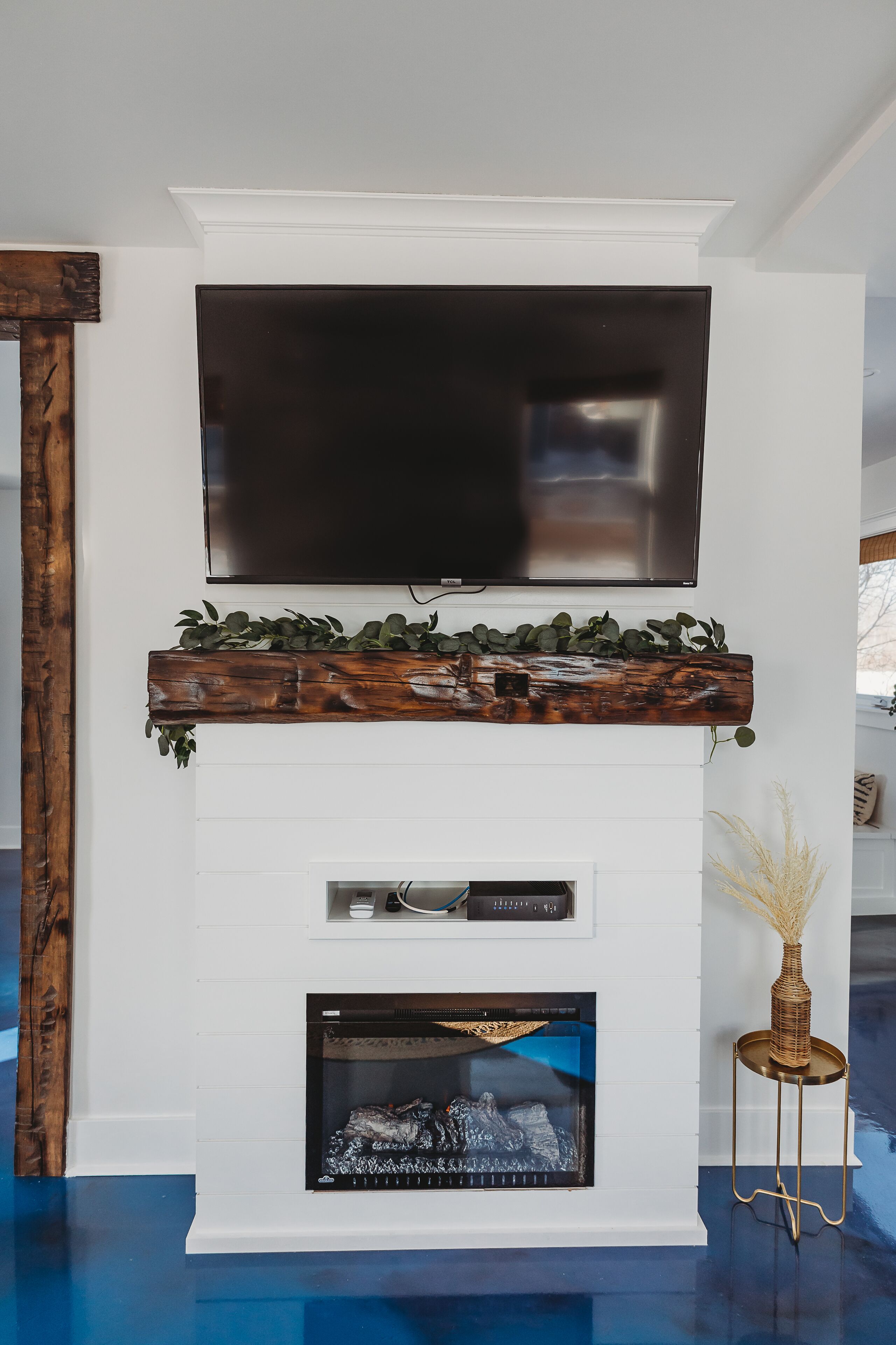 A modern living room with a flat-screen TV mounted above a wooden mantelpiece with greenery, a fireplace below, and a small side table with a vase.