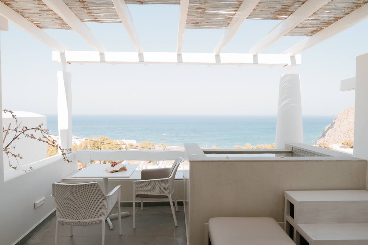 Un balcón con vistas al mar, muebles blancos y una estructura de pérgola.