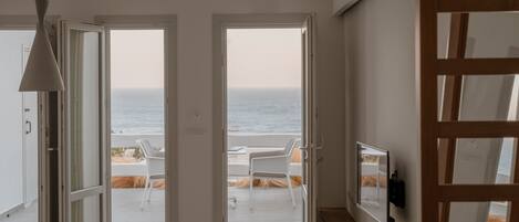 A room with a view of the sea through open doors. There is a white chair and a table outside.