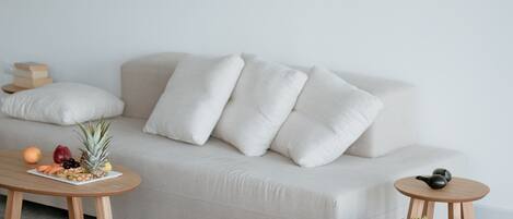 A modern living room with a beige sofa, white cushions, a wooden coffee table with fruit, a small round table, and two white poufs.
