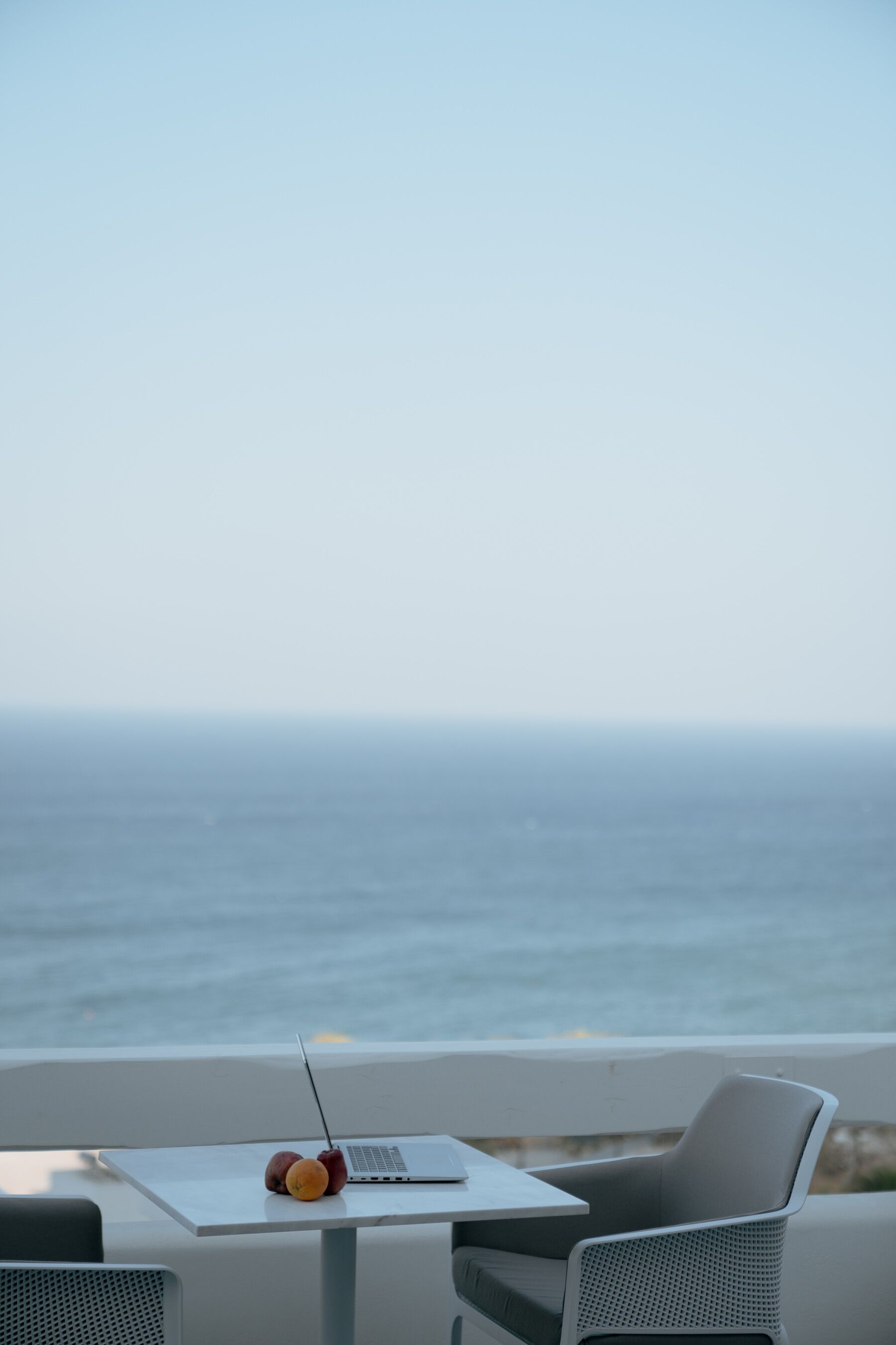 Una laptop sobre una mesa con dos manzanas y una taza, con vista a una playa y el océano al fondo.