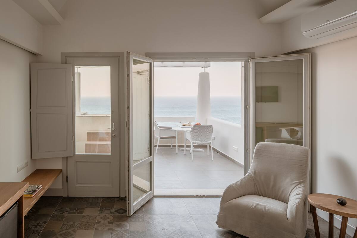 Una habitación con vista al mar, una silla blanca, una mesita pequeña y una puerta que da a un área de estar al aire libre.