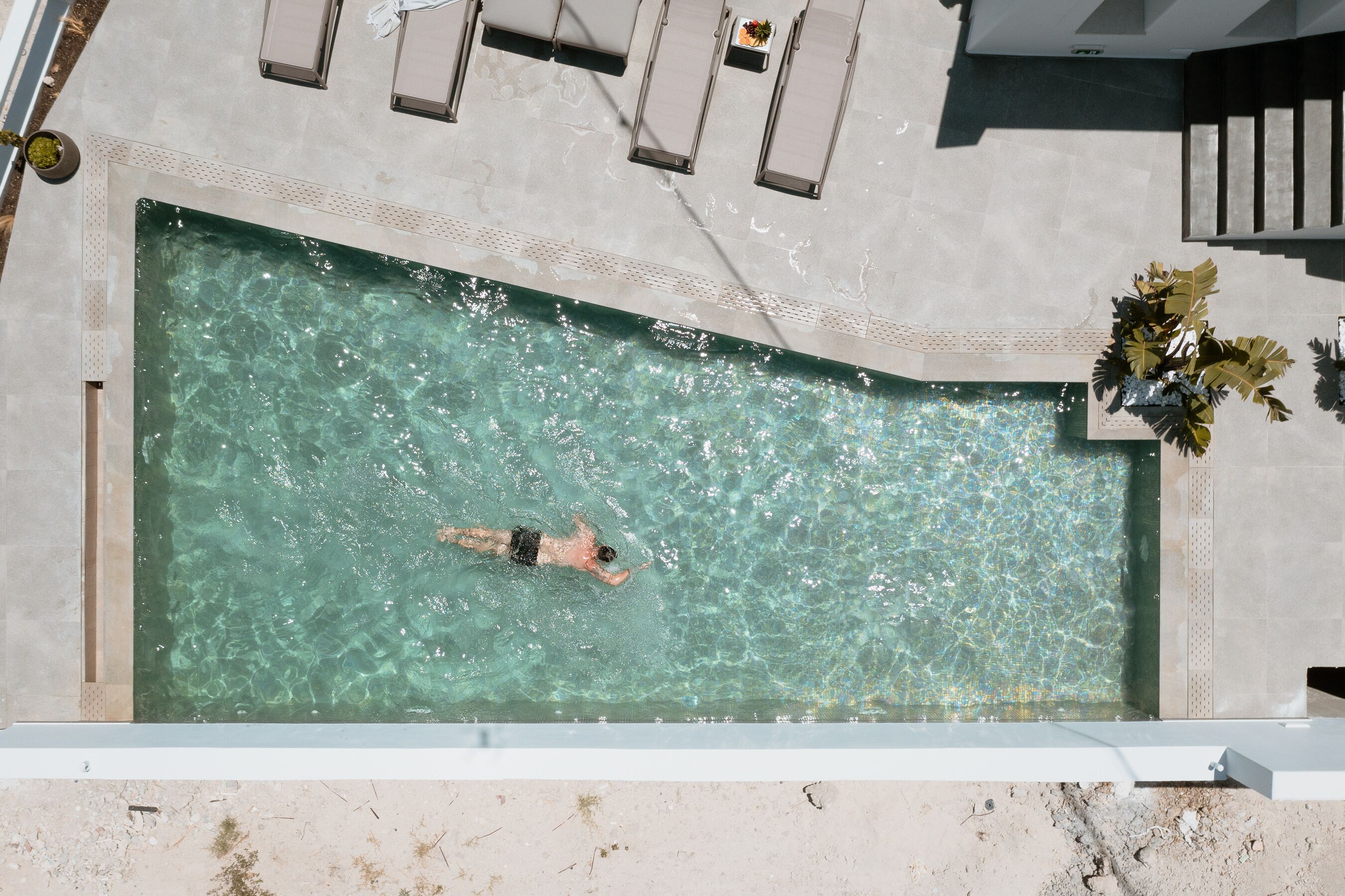 A swimming pool with a person swimming, surrounded by a tiled deck and lounge chairs.