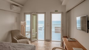 A hotel room with a bed, a desk, a chair, a lamp, and a TV. There is a balcony with a view of the sea.