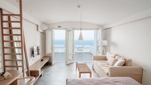 A modern hotel room with a bed, a desk, a chair, a sofa, and a view of the sea.