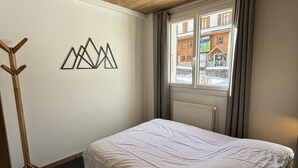 A bedroom with a wooden ceiling, a bed, a coat rack, a window with a view of buildings, and a wall art piece. - 350m from ski slopes, 2nd floor, sauna, terrace, ski locker, 41m², Valloire (Valloire)