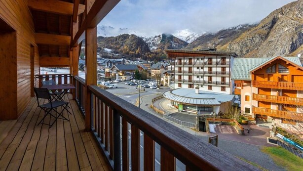 A wooden balcony with a table and chairs, overlooking a town with buildings and mountains in the background.