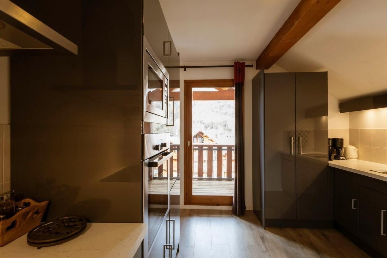 Una cucina moderna con mobili scuri, un piano di lavoro in legno e vista su un patio in legno e una casa attraverso una porta vetrata.