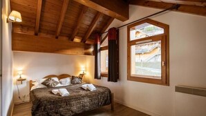 A bedroom with a wooden ceiling, a large window with curtains, a bed with a dark patterned bedspread, and two bedside lamps.