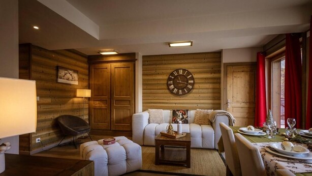 A cozy living room with a sofa, ottoman, and coffee table, wooden walls, and a large wall clock.
