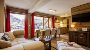 A spacious living room with a large sofa, a dining area with a table set for four, and a kitchen with wooden cabinets and a TV mounted on the wall.