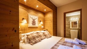 Una camera da letto in stile baita con un letto, biancheria a quadri, un’opera d’arte raffigurante una testa di cervo e un bagno visibile attraverso una porta aperta.