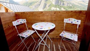 Un balcón de madera con dos sillas plegables blancas y una mesita redonda, con vistas a un paisaje montañoso.