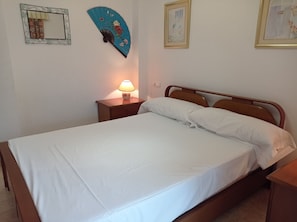 Uma cama bem arrumada com lençóis brancos, uma luminária de cabeceira e um ventilador decorativo fixado na parede.