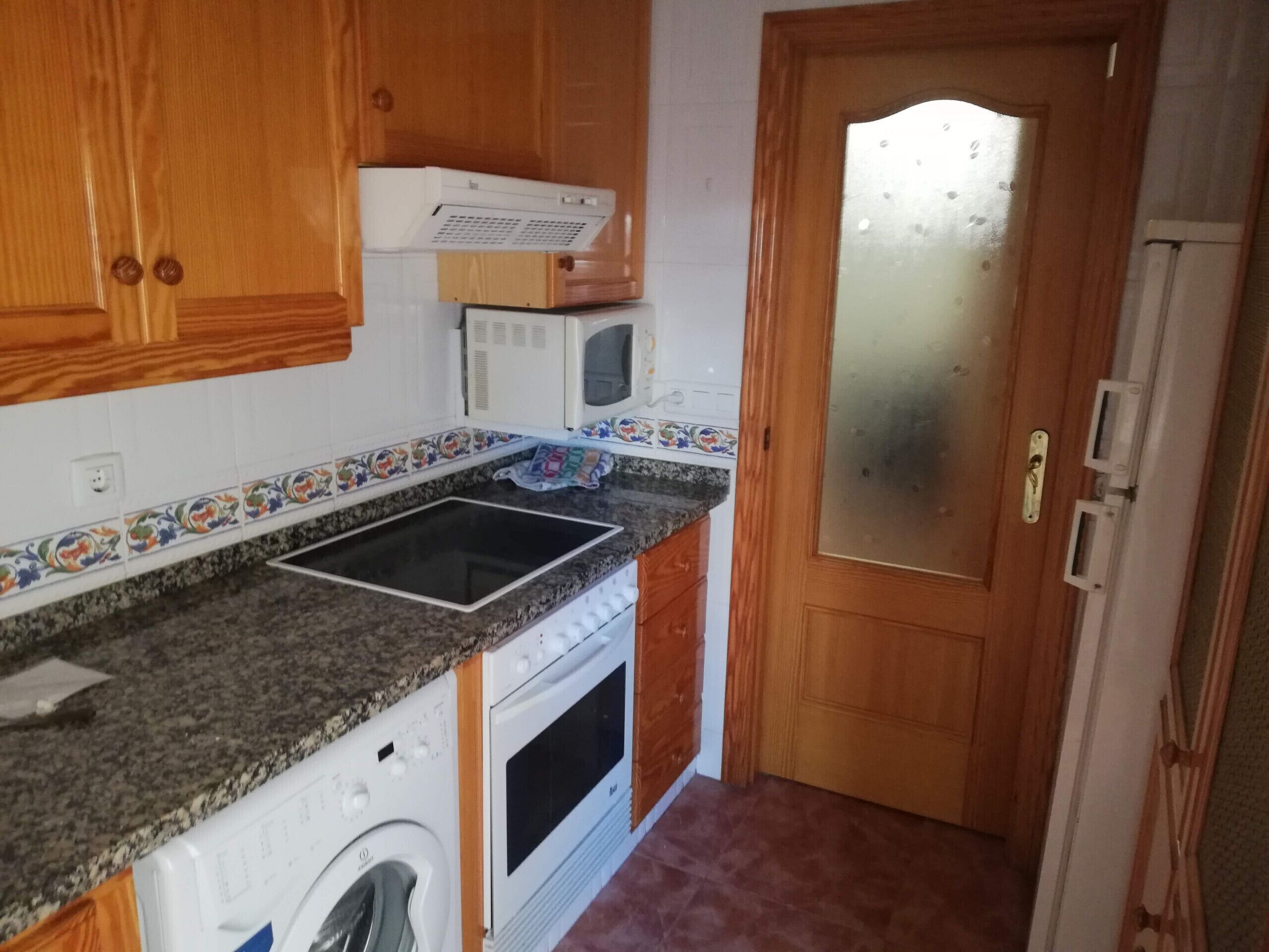 A compact kitchen with a built-in microwave, a washing machine, and a wooden door with a frosted glass panel.