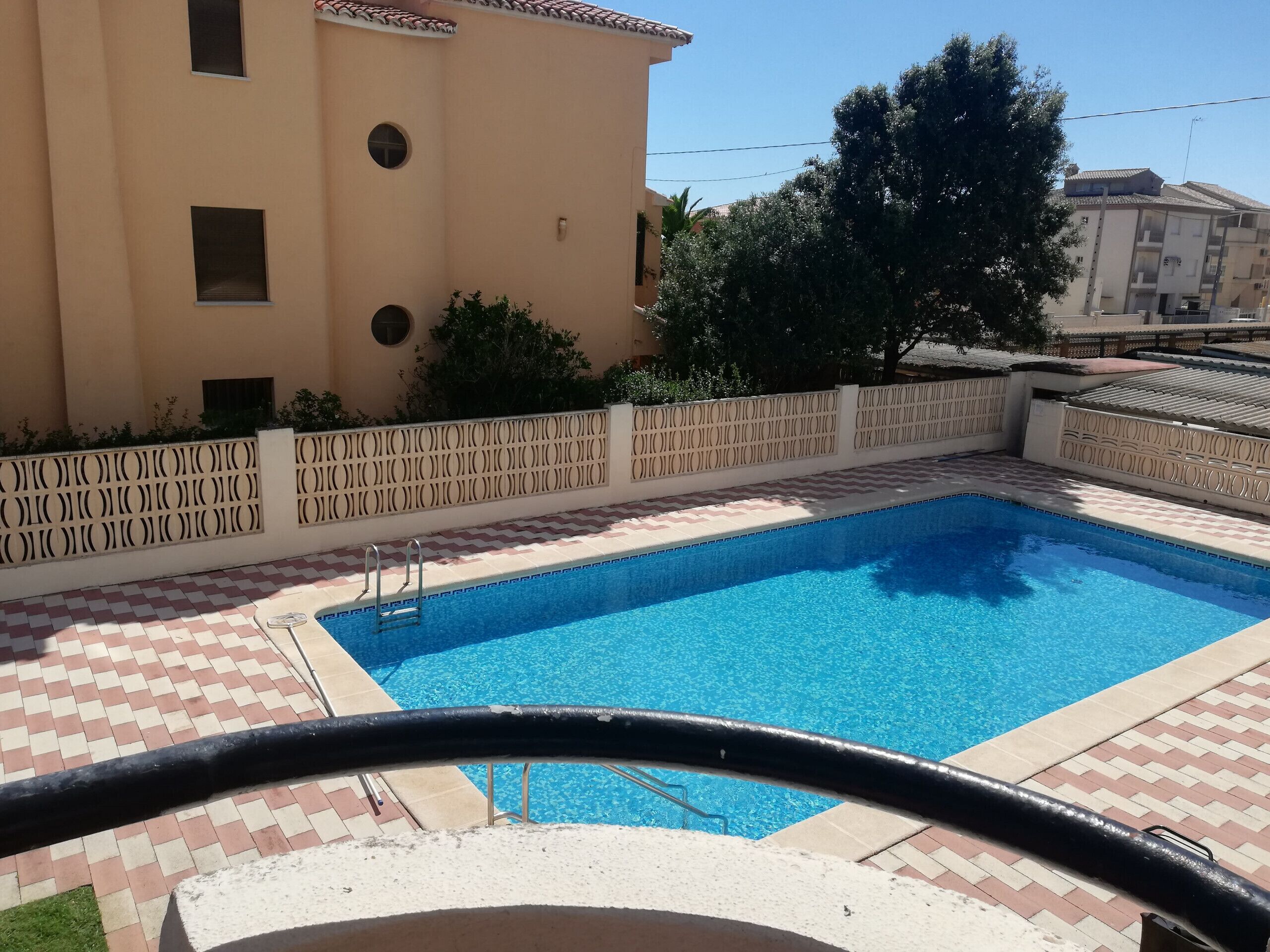 En swimmingpool med fliseterrasse, et hvidt hegn og en bygning med beige facade.