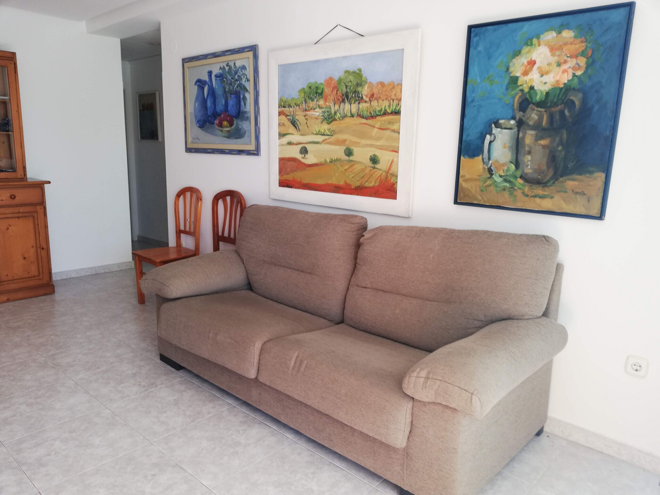 Een woonkamer met een beige bank, houten stoelen en drie schilderijen aan de muur.