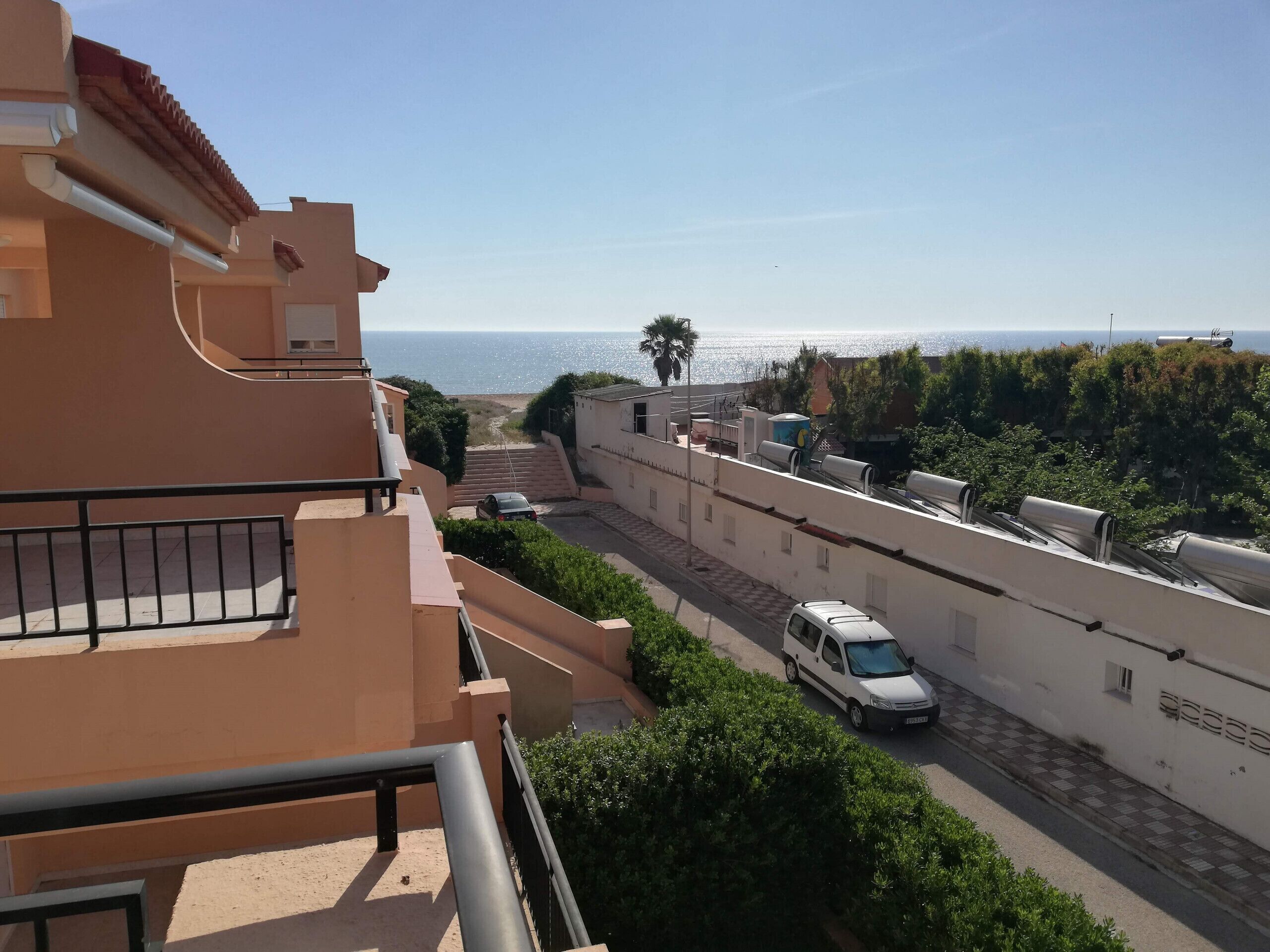 Eine Wohnstraße mit Blick auf das Meer, ein weißer Transporter parkt am Straßenrand und ein Balkon mit Geländer.