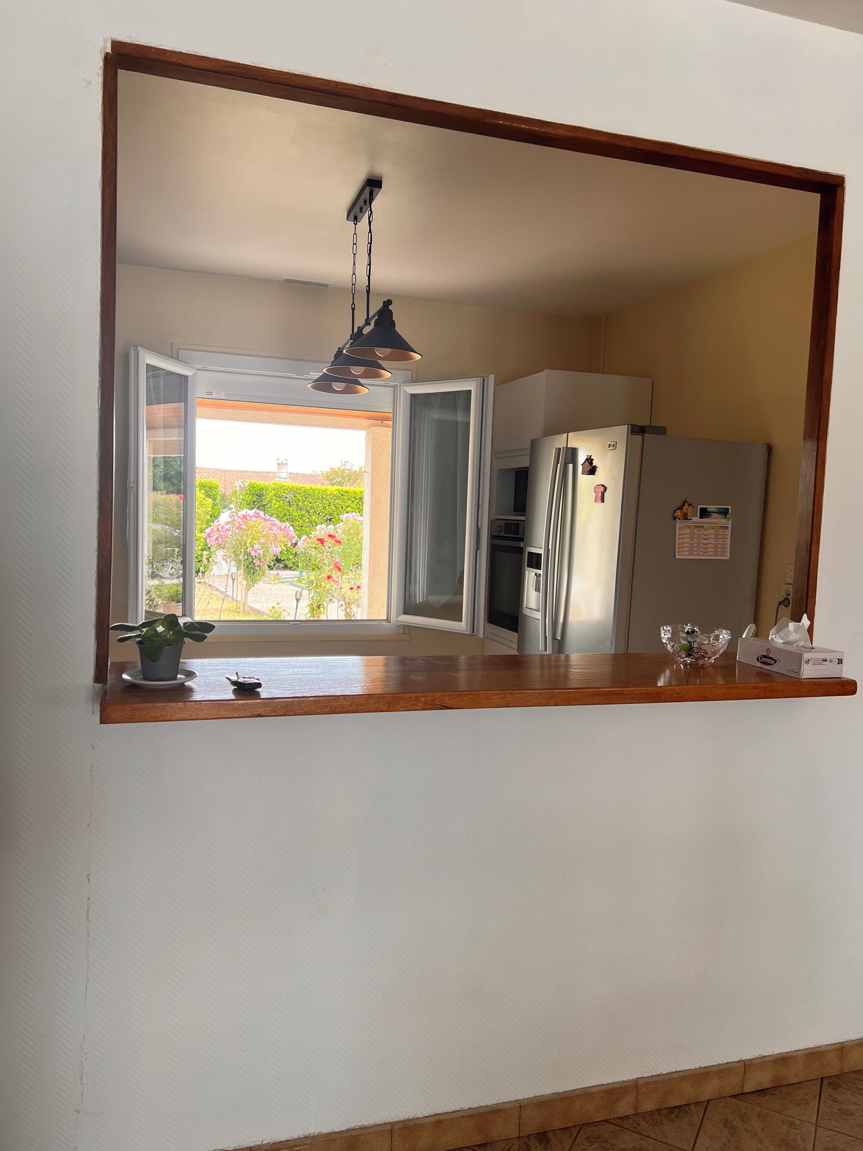 A kitchen with a large mirror, a hanging light fixture, a refrigerator, and a window with a view of a garden.