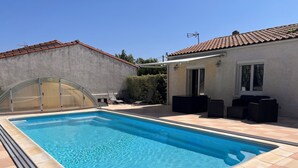 A rectangular swimming pool with a tiled deck, surrounded by a covered area with chairs and a small table. - Villa with private pool (Saint-Froult)