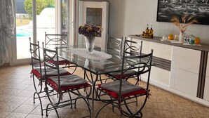 A dining area with a glass table and metal chairs, a vase with flowers, a painting on the wall, and a view of the outside. - Villa with private pool (Saint-Froult)