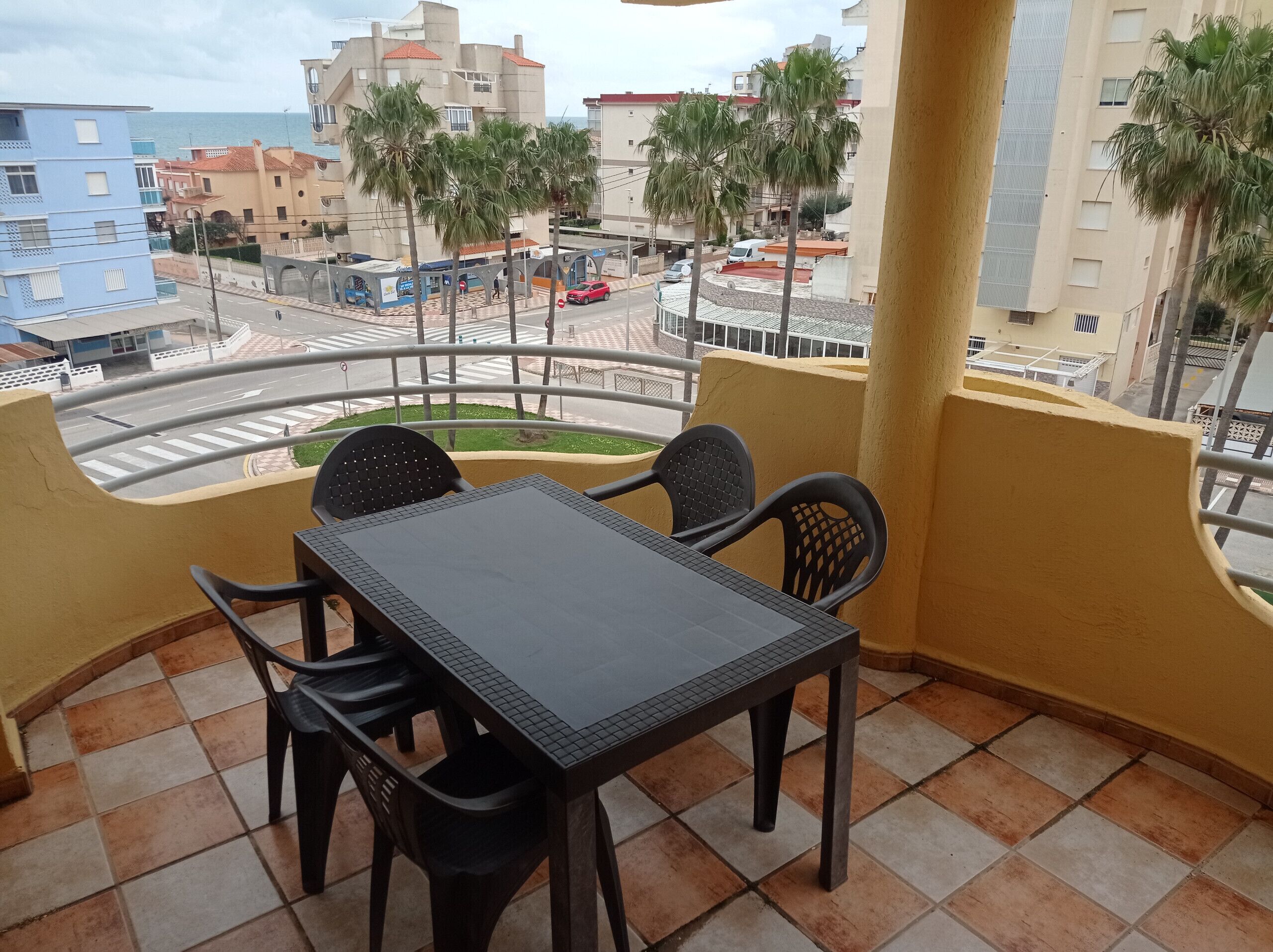 A balcony with a table and chairs, overlooking a street with buildings and palm trees.