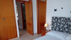 Un dormitorio con cama, mesita de noche con lámpara, puertas de madera y un baño visible a través de un acceso abierto.
