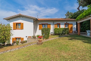 A two-story house with a tiled roof, white walls, and wooden shutters. There is a paved pathway leading to the entrance, and a small garden area with potted plants and flowers.