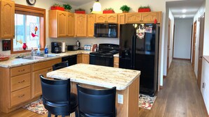 A modern kitchen with wooden cabinets, a black refrigerator, a microwave, and a coffee maker. There is a window with a decorative view, and a chandelier hangs from the ceiling. - Cheerful cottage w/ fireplace. Great location for exploring the Lake Pepin area! (Pepin)