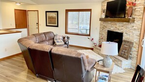 A modern living room with a brown leather sectional sofa, a fireplace, a flat-screen TV, and a stone wall. - Cheerful cottage w/ fireplace. Great location for exploring the Lake Pepin area! (Pepin)